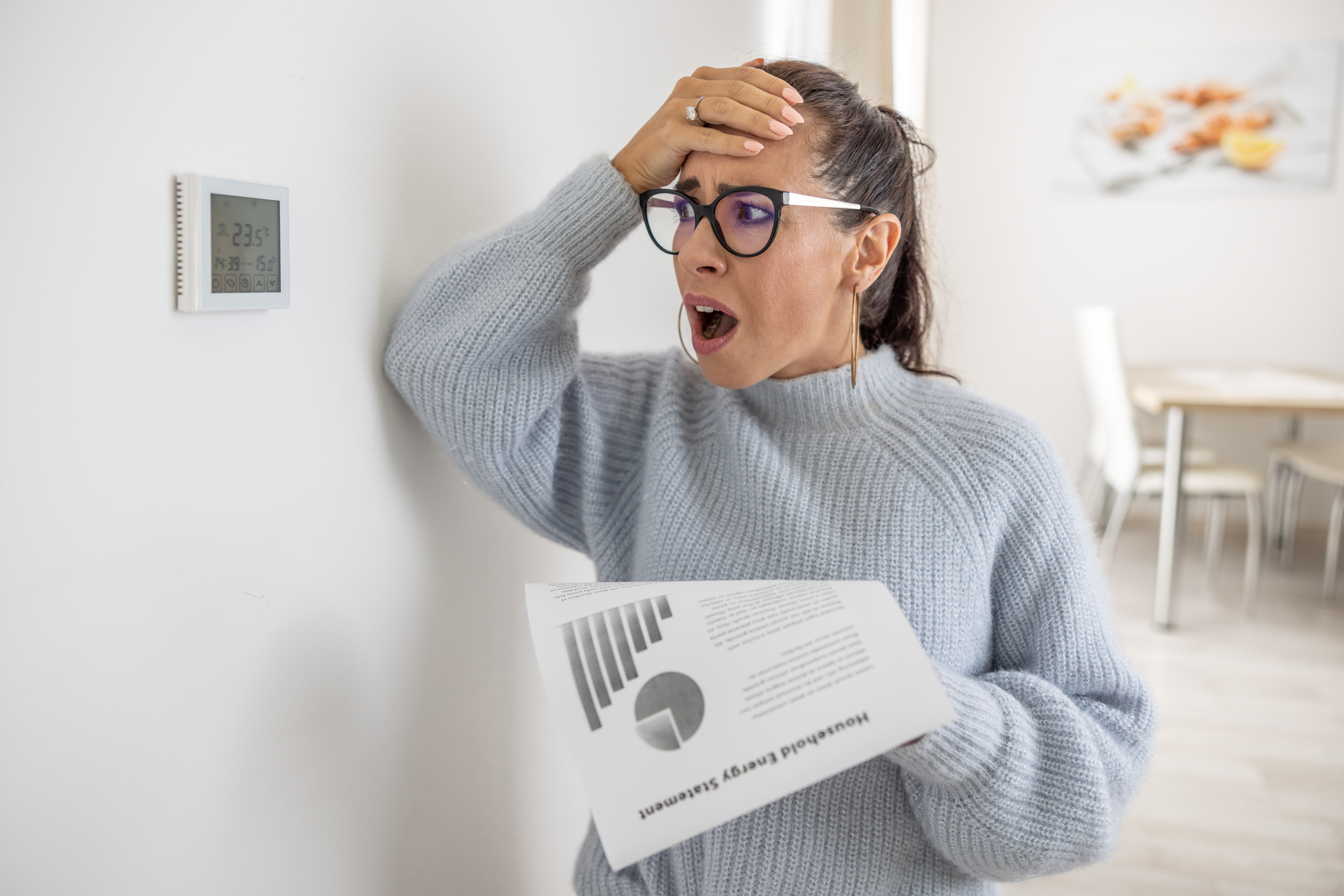 Woman holding high winter energy bill while looking at thermostat